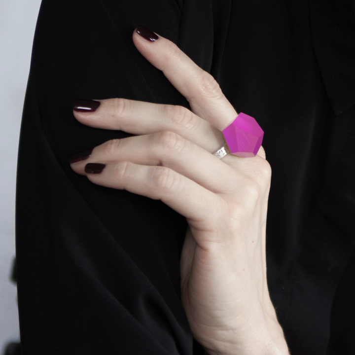 A hand with dark maroon nail polish is wearing a large, geometric-shaped, bright pink Fruit Bijoux Ring Top VU Crystals Magenta Pink atop a sterling silver band. The person is dressed in a black garment.