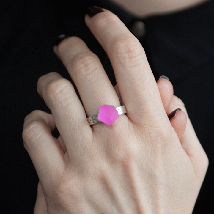 A close-up of a hand wearing the Fruit Bijoux Ring Top VU Crystals Magenta Pink by Fruit Bijoux. The person's nails are painted dark, and the background is a dark fabric, possibly part of their clothing.