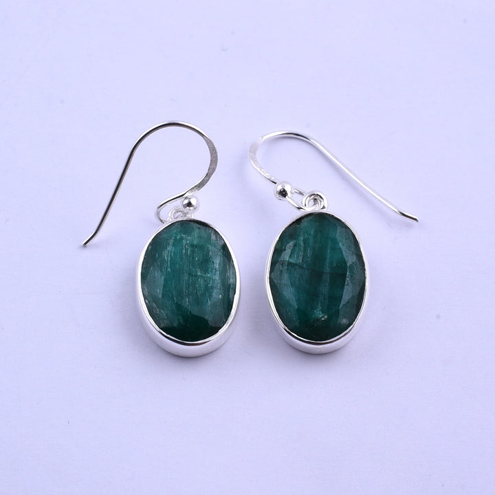 A pair of sterling silver hook earrings featuring oval-shaped, faceted green gemstones. The Simple Silver & Rough Emerald Oval Earrings by Watermelon Tropical showcase a contemporary design with the green stones set in a sleek silver bezel. The plain white background highlights the elegance of the jewelry.