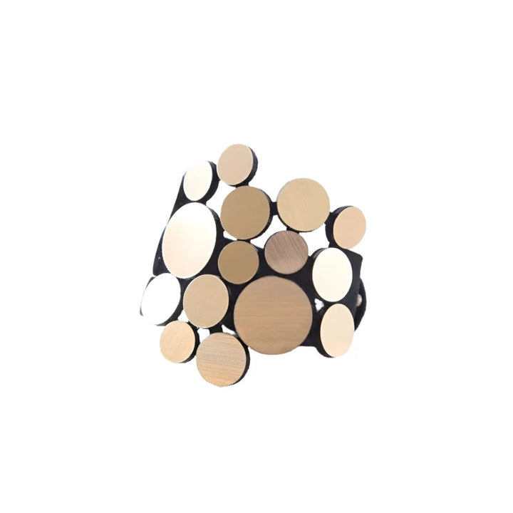 The Abstraction Fizz Bracelet by Iskin Sisters is a stylish accessory featuring an array of circular gold-colored discs of varying sizes, all elegantly arranged on a black band. It is displayed on a light-colored, irregularly shaped stone surface with a soft, neutral background.