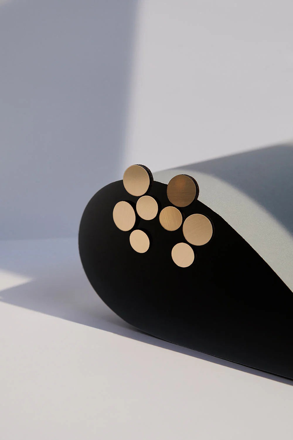 A minimalist photograph showcases the Abstraction Gold Earrings by Iskin Sisters—various sized golden circular discs reminiscent of lightweight statement earrings, arranged in a scattered pattern on a curved black and light gray surface, set against a neutral background. The image captures a play of light and shadow, emphasizing the metallic textures.