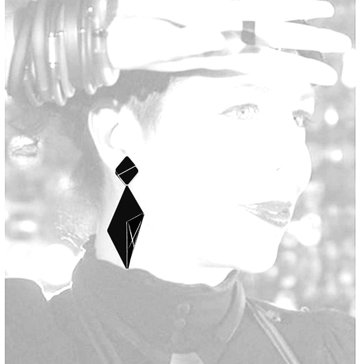 A high-contrast, black-and-white image features a person wearing minimalist Alicia Earrings by Alex + Svet, intricately shaped like modern sculptures. The person, partially obscured by overexposure, holds a hand to their forehead, adorned with multiple black bracelets.