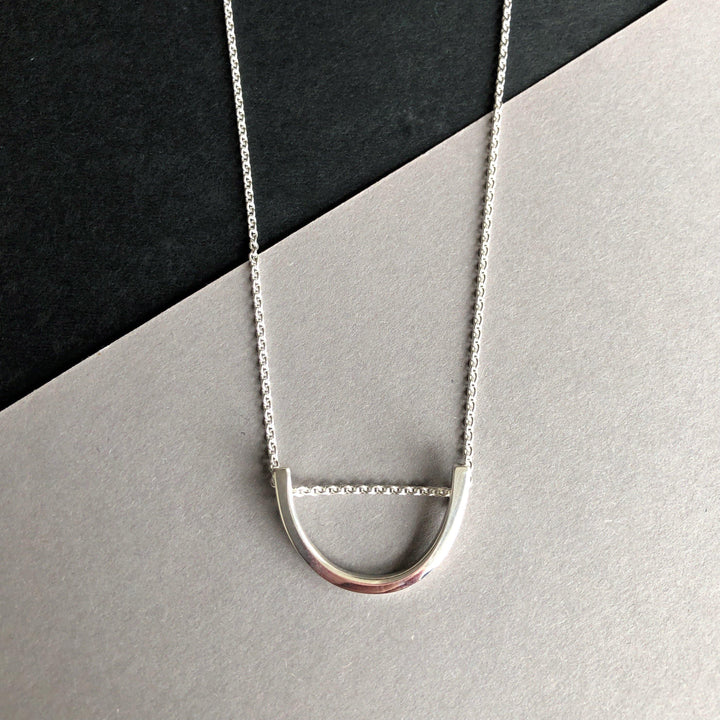 A handmade Silver Curve Pendant by Angela O'Keefe displayed against a dual-tone black and gray background. The delicate chain and minimalist design of the sterling silver bar create a sleek, modern look inspired by Ireland's craftsmanship.