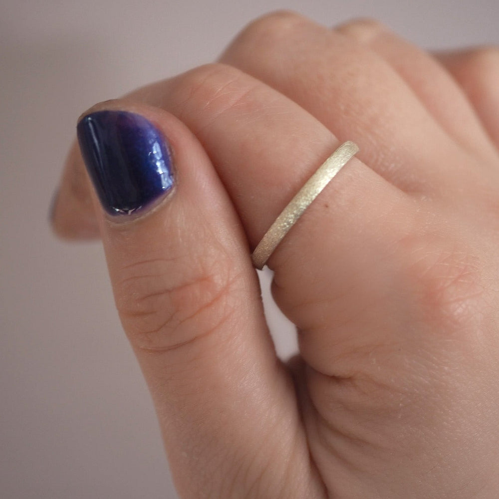 A close-up of a hand with a plain, thin gold ring on the ring finger. The nails, painted with glossy dark purple nail polish, contrast beautifully against the blurred, neutral background. Next to the gold ring is the Ellipse Stacking Ring Silver by Aud.