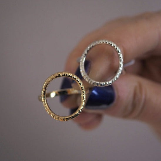 Close-up of a hand with dark purple nail polish holding two metal rings. One is a unique, handmade gold ring, and the other is an Aud Into The Light Ring Silver, both featuring textured, serrated designs around their circular shapes. The background is neutral and blurred.