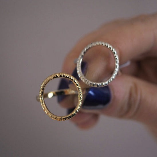 A hand with navy blue painted nails holds two rings side by side. Both rings feature large, textured circular designs. The gold-colored one is the **Into The Light Ring 9ct Yellow Gold** by **Aud**, while the other is silver-colored. The background is blurred in a soft beige tone reminiscent of the view from Hook Lighthouse.