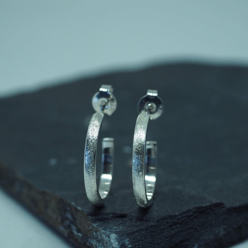 A pair of Ellipse Small Hoops Silver by Aud is displayed on a dark, slate-like surface. The background is softly blurred, highlighting the earrings' metallic shine and intricate details. Perfect for everyday wear, these handmade jewelry pieces feature latch-back closures for secure wear.