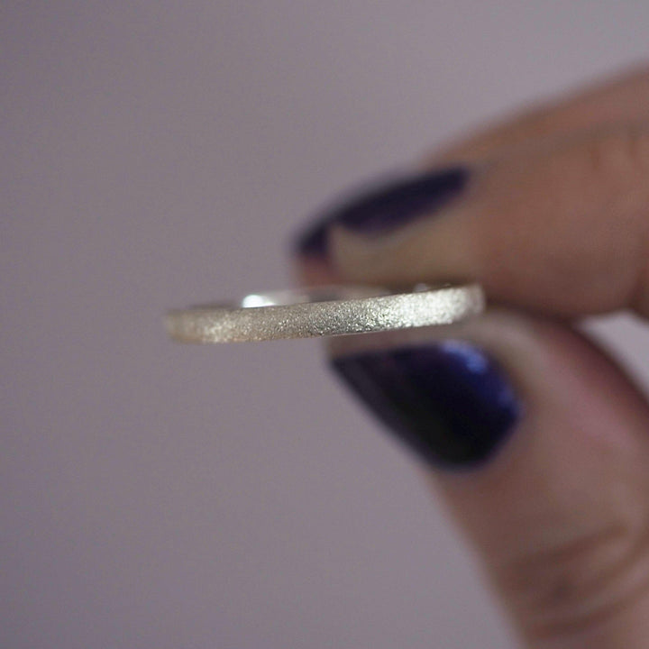 A person with dark purple nail polish holds a thin, sterling silver ring between their thumb and index finger. The background is blurred, drawing attention to the frosted texture of the Ellipse Stacking Ring Silver by Aud.