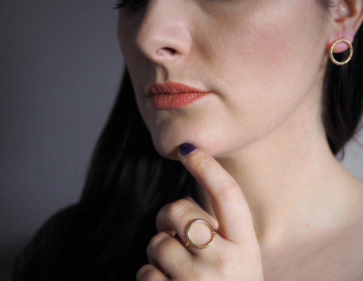 Close-up of a person with long dark hair, wearing Aud Into The Light Circle Earrings 9ct Yellow Gold and a matching ring. They are gently touching their chin with their index finger. The background is neutral.