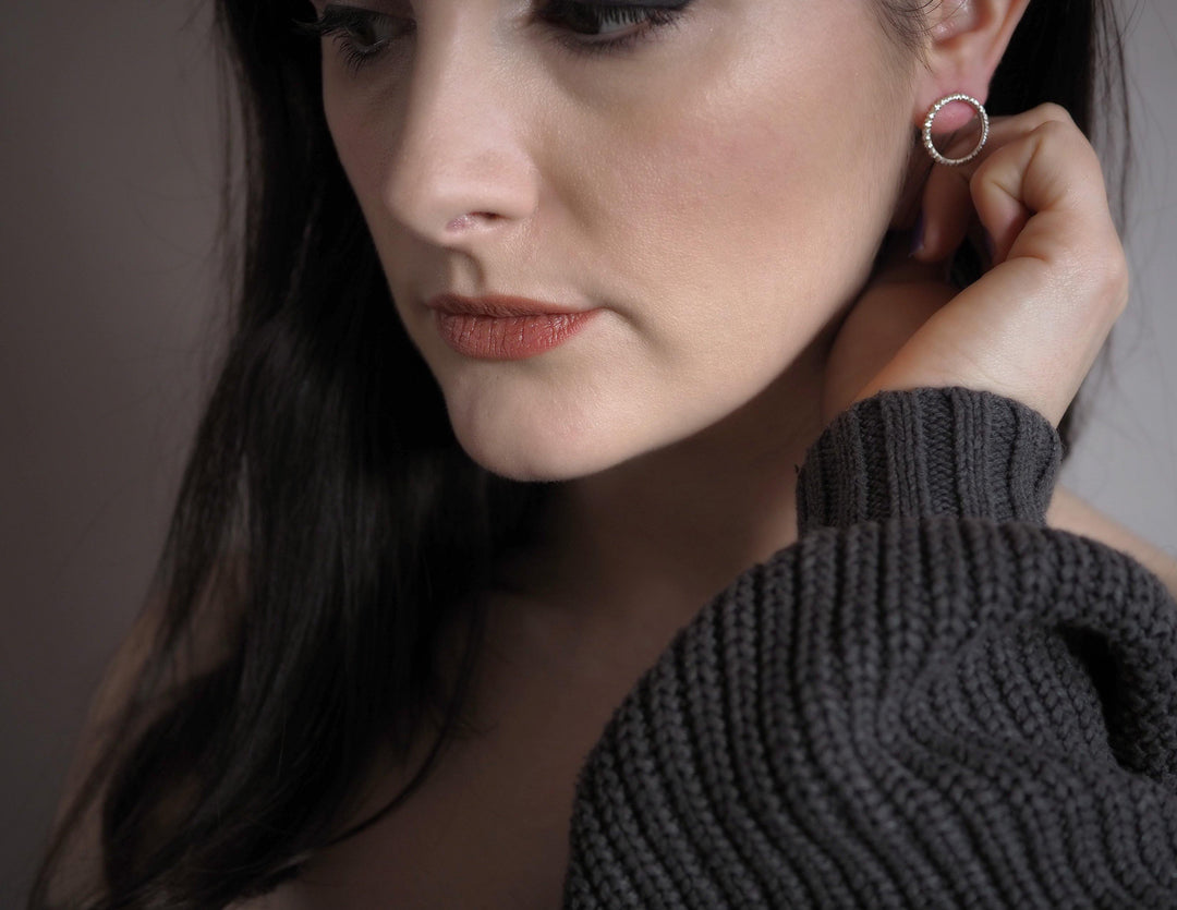 A close-up of a person with long dark hair and light skin wearing a dark gray knitted sweater. They are looking down, their hand touching an Into The Light Circle Earrings Silver by Aud. The texture detail of the knitted fabric is prominent, complementing their neutral, contemplative expression and subtle makeup.