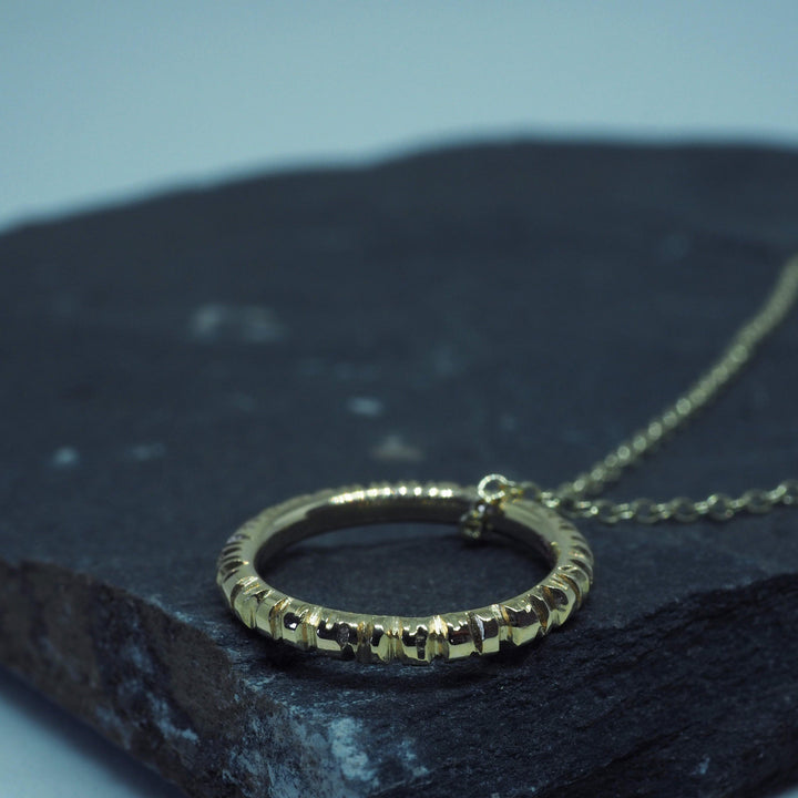 An Into The Light Circle Pendant 9ct Yellow Gold by Aud hangs from a delicate chain, showcased on a dark, textured stone surface. This unique necklace's intricate design contrasts with the smooth background, highlighting its elegant craftsmanship in handmade jewelry.