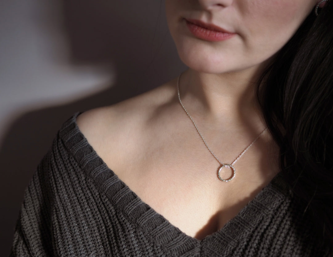 Close-up of a person wearing a ribbed, knitted sweater and a delicate, handmade "Into The Light Circle Pendant Silver" necklace crafted from sterling silver by Aud. The image focuses on the neckline and the unique jewelry, highlighting the subtle texture of the sweater and the intricate design of the necklace.