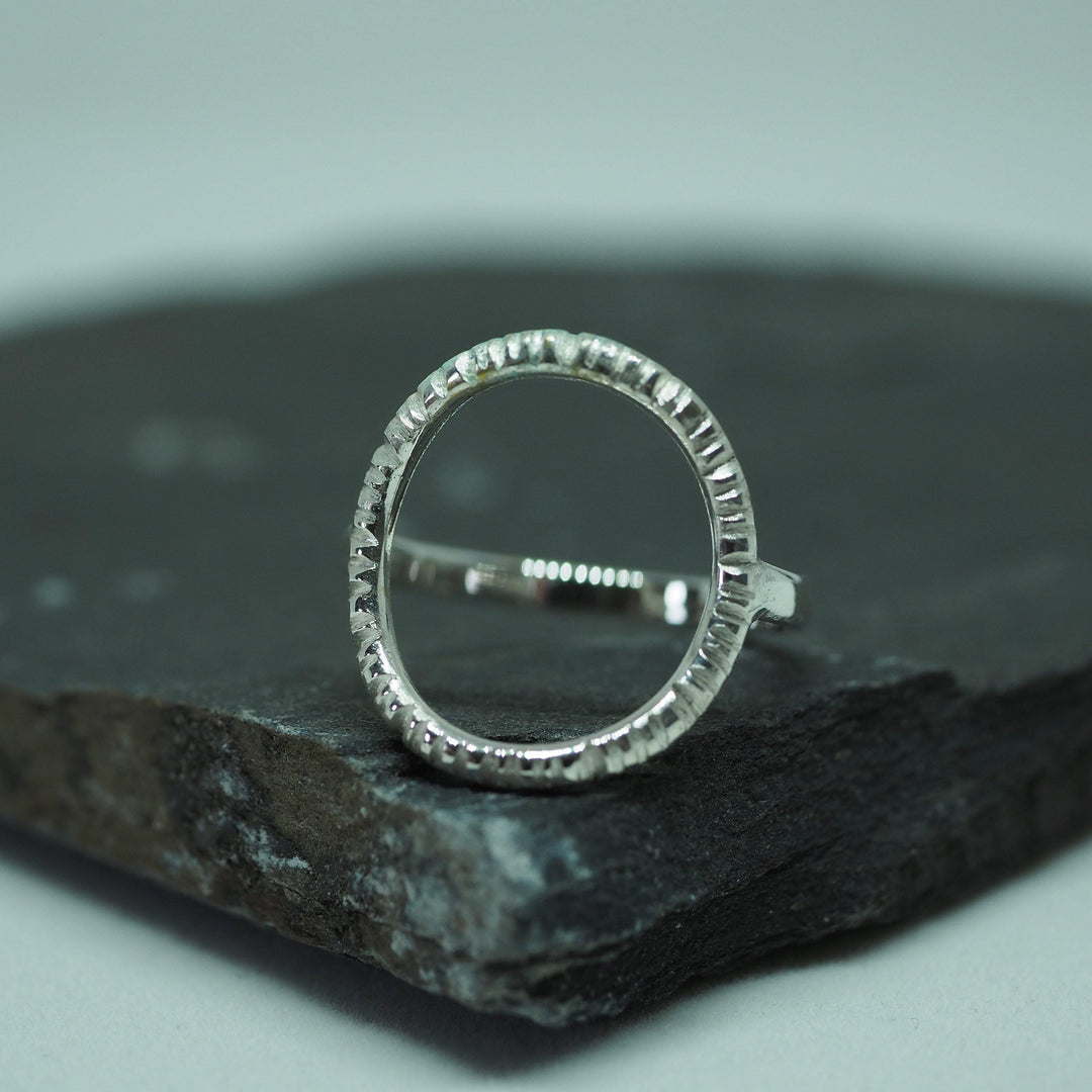 A unique, handmade sterling silver ring with a textured circular band is placed on a dark, rectangular stone surface. The Into The Light Ring Silver by Aud appears shiny and intricately crafted, with a modern and minimalist design. The background is a soft gradient of light grey.