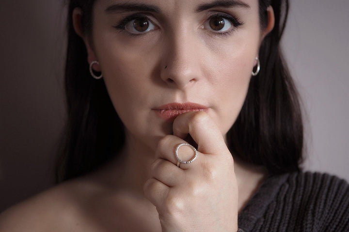 A woman with long dark hair and Into The Light Circle Earrings Silver by Aud is looking directly at the camera with a thoughtful expression. She is wearing a dark, off-the-shoulder sweater and has a ring on her index finger, which has intricate texture detail, resting against her lips.