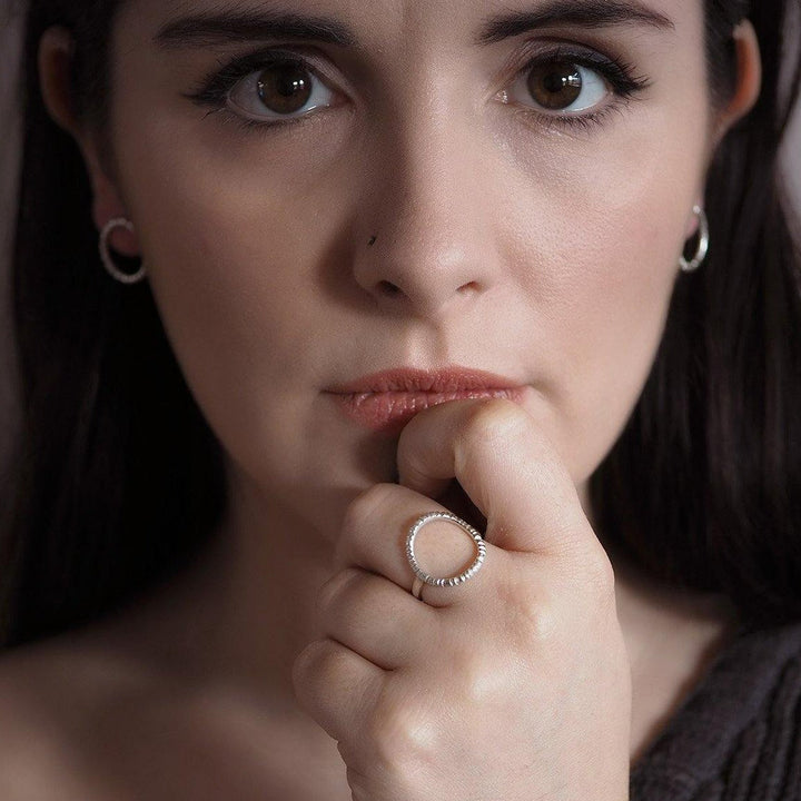 A close-up portrait of a woman with dark hair and brown eyes, gazing directly at the camera. She wears small hoop earrings and showcases an Aud Into The Light Ring Silver on her right hand, which she holds near her lips. The background is softly blurred.