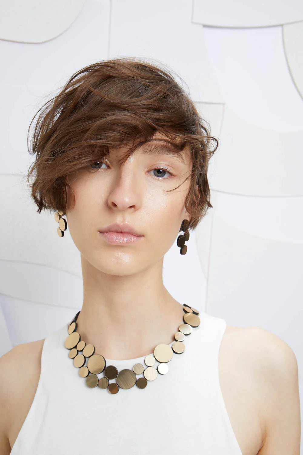 An individual with short, tousled brown hair and minimal makeup is wearing a white sleeveless top. They have a matching necklace and earrings set from Iskin Sisters' collection, featuring circular metallic pieces in various sizes. The necklace is the Abstraction Fizz Gold Necklace from Iskin Sisters. The background features a white, abstract, geometric pattern.