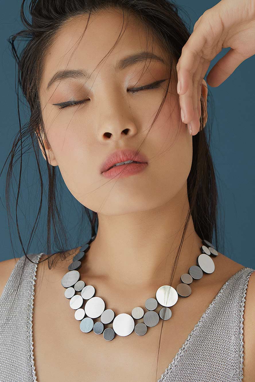 A woman with long dark hair poses with her eyes closed in front of a blue background. She wears a light gray, sleeveless top and an Abstraction Fizz Silver Necklace from Iskin Sisters, featuring variously sized round metallic discs. One hand is raised near her forehead, and her expression is calm and serene.