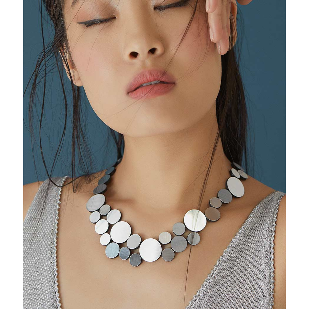 A woman with long dark hair and closed eyes wears a sleeveless knit top and the Abstraction Fizz Silver Necklace by Iskin Sisters, posing against a blue background.