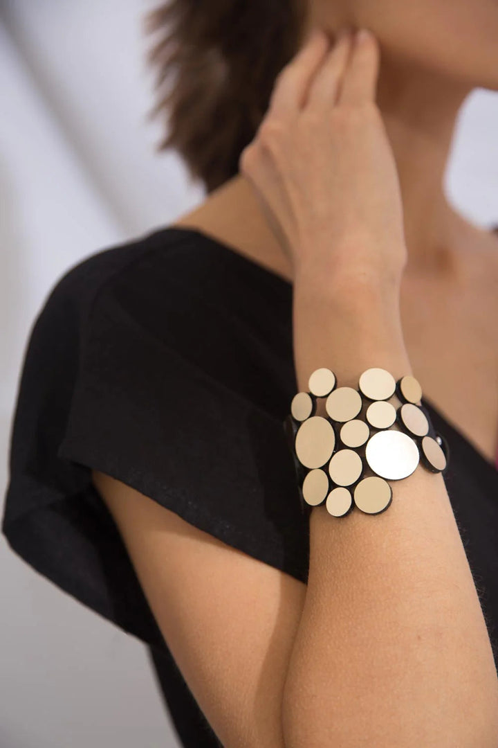 A person wearing a black top showcases the Abstraction Fizz Bracelet by Iskin Sisters on their wrist. The bracelet features multiple overlapping, differently sized circular disks in a metallic finish. The person's hand is gently touching their neck, and the background is blurred.