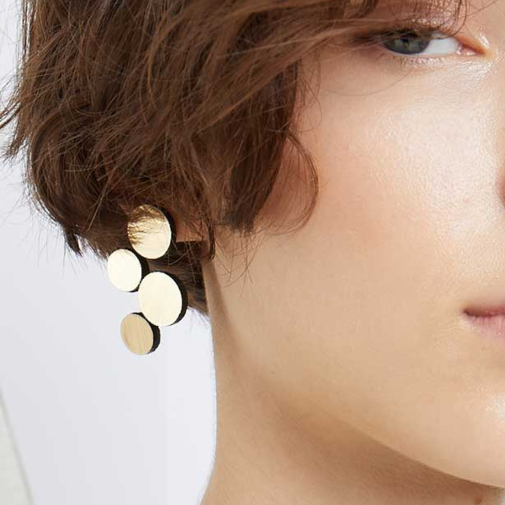 Close-up of a person with short brown hair wearing Iskin Sisters' Abstraction Gold Earrings—geometric accessories featuring three shiny, round discs. Only part of their face is visible in the image.