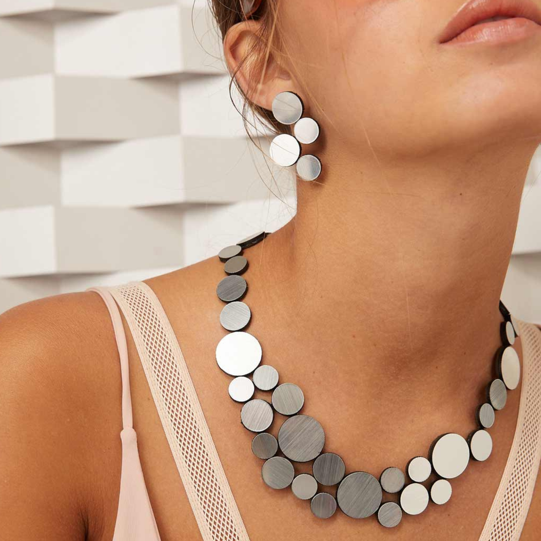 A woman models the Iskin Sisters Abstraction Silver Earrings—bold, lightweight geometric jewelry in silver and black—paired with a sleeveless light pink top against a white geometric background.