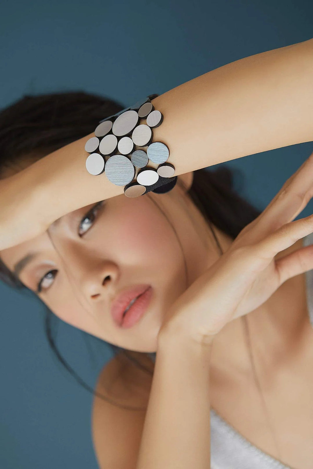 A person with long hair holds their arm across their forehead, showcasing an Abstraction Fizz Bracelet by Iskin Sisters, made of overlapping round metal discs. They are gazing towards the camera, with a blue background behind them, epitomizing the artistic design often crafted by talented female artisans.