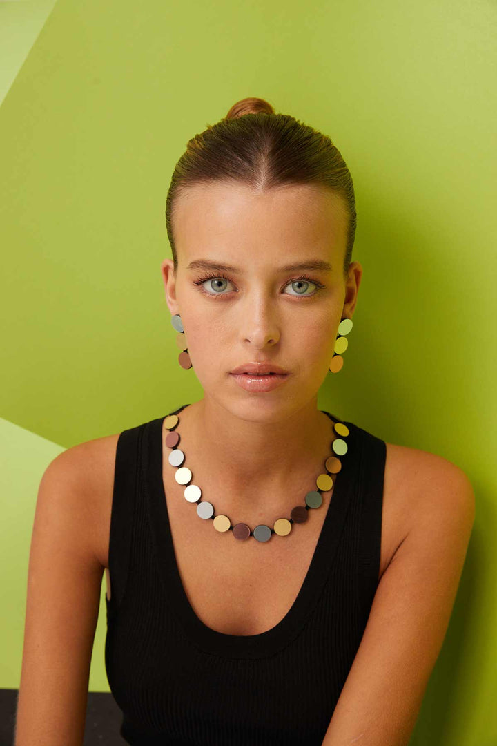 A person with light blue eyes and hair tied back in a bun is wearing a black sleeveless top. They have large, circular earrings and are adorned with the Abstraction Silver, Gold & Rose Gold Necklace by Iskin Sisters. The background features a green geometric pattern.