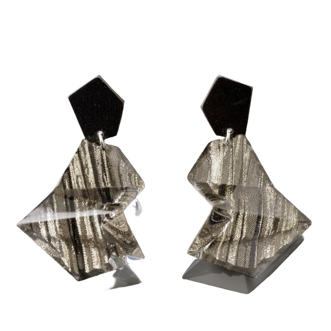 A pair of Acrylic Black & Silver Earrings by MX.design. Each earring has a black, irregular hexagonal top piece connected to a metallic, crumpled, and triangular base with reflective ridges. The background is plain gray.