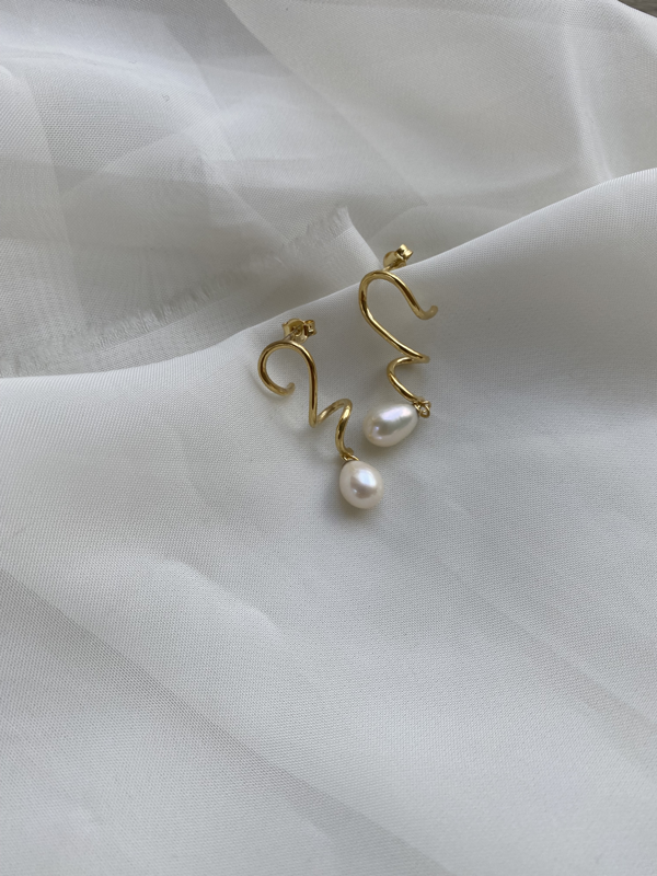 A pair of Bonvo Agne Pearl Earrings, 18ct gold plated spiral earrings each with a single dangling white pearl drop, sits on a soft, sheer white fabric. The elegant design contrasts beautifully with the delicate background.