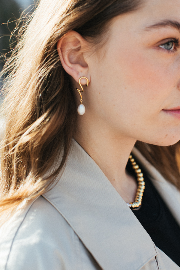 Close-up of a person with long brown hair wearing a beige trench coat, a gold necklace, and Bonvo's Agne Pearl Earrings. The photo captures a side view focused on the head and upper shoulder area, showcasing the earring’s 18ct gold plating over solid 925 sterling silver.