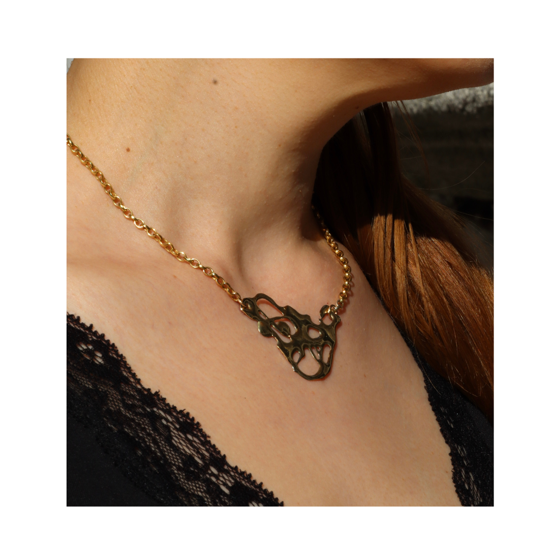 A person with reddish-brown hair wears the Alchemy Necklace by Madeleine Sculptural Jewellery, a handmade gold chain with an abstract pendant, styled over a black lace-trimmed top.