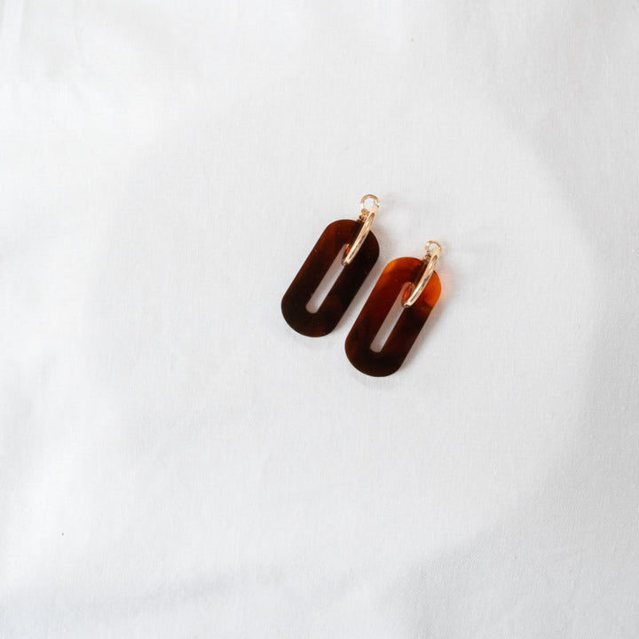 A pair of Amber Oval Acrylic Earrings by Studio Nok Nok with a tortoiseshell pattern linked to small gold hoops, crafted from recycled acrylic and placed on a white fabric background.