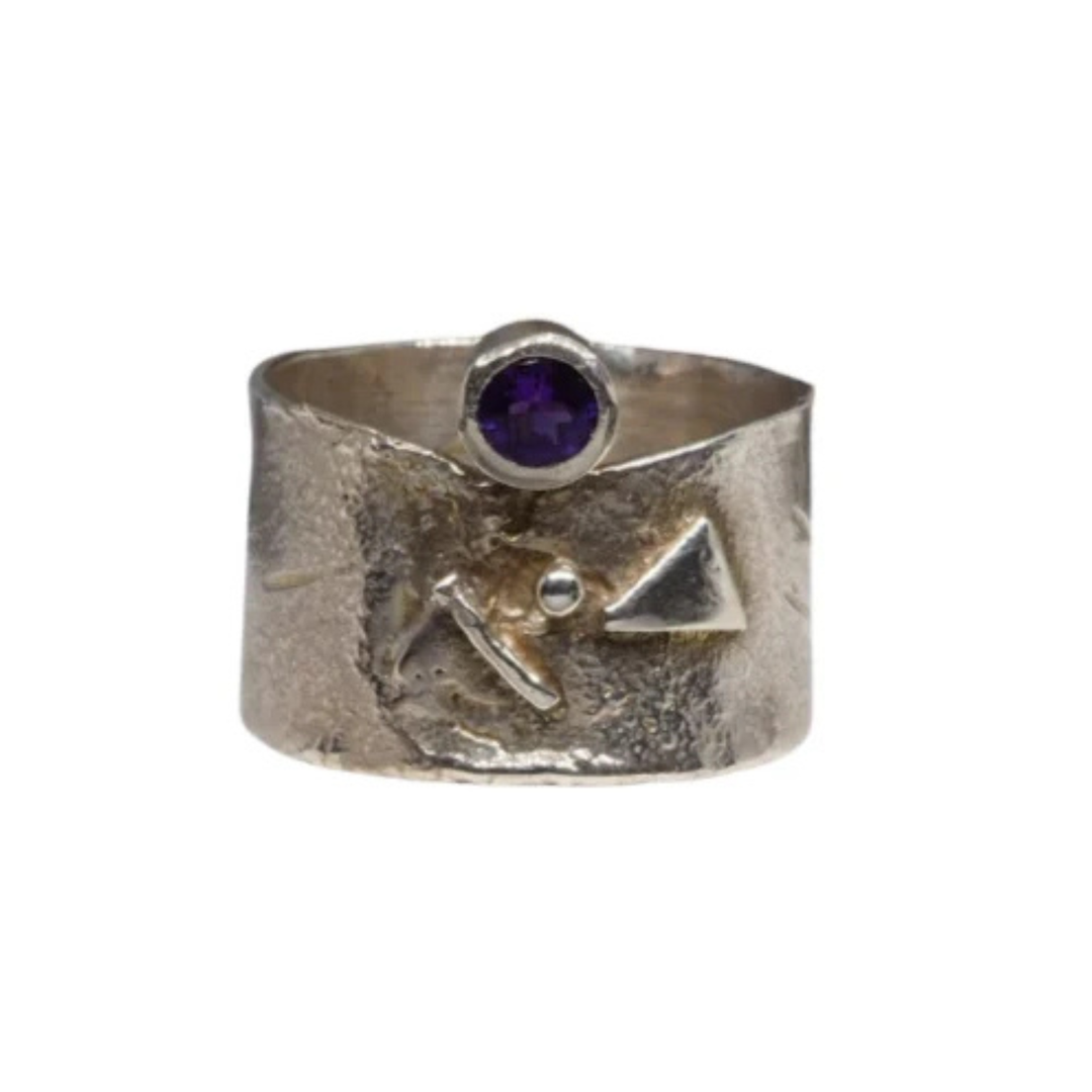A wide sterling silver ring with a textured surface and an abstract design featuring an amethyst gemstone set into a small circular setting atop the band. The Amethyst Guardian Ring by Caroline Stokesberry-lee includes raised triangular and round shapes, adding to its unique charm.