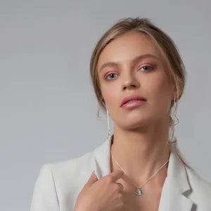 A woman with light skin and blonde hair wears a light blazer and delicate Ammil Long Earrings by Inner Island, posing confidently against a plain gray background.