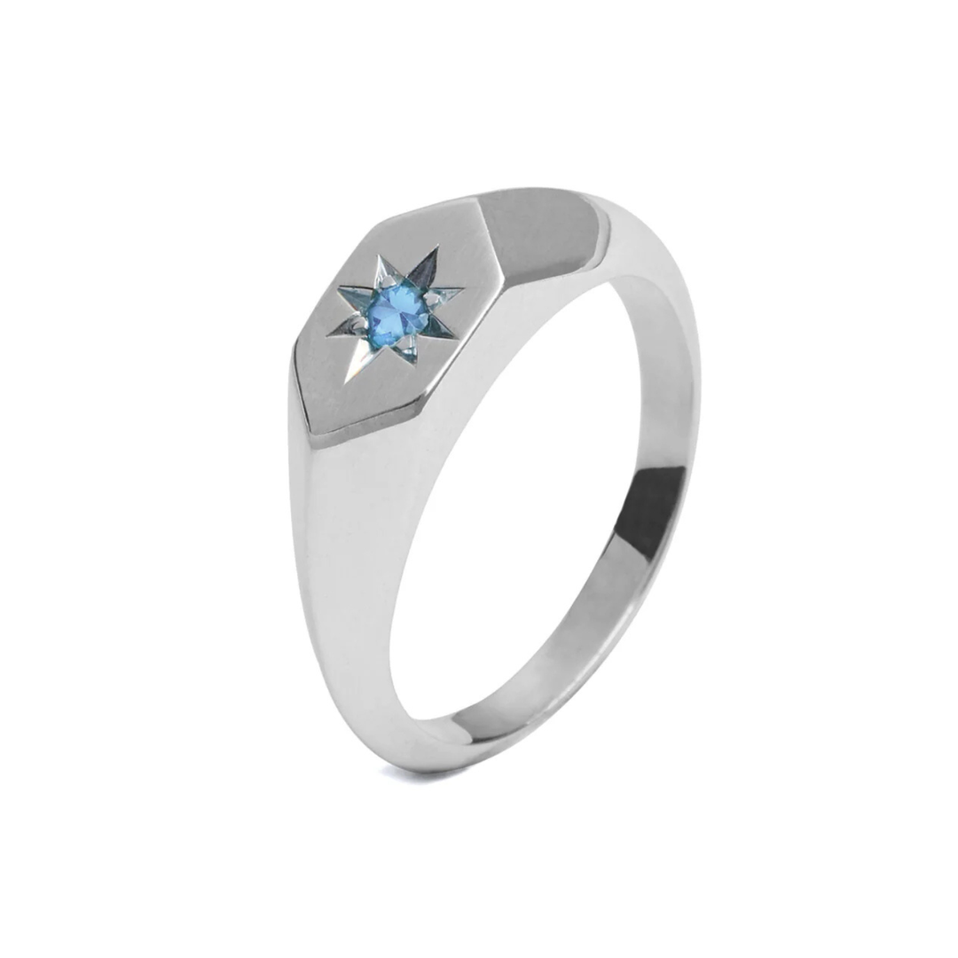 A Cosmic Boulevard Starlight Blue Zircon Birthstone Signet Ring with a hexagonal top featuring a small blue zircon gemstone embedded in the center, surrounded by an engraved starburst pattern, perfect for celebrating December birthdays. The ring has a smooth, polished finish.
