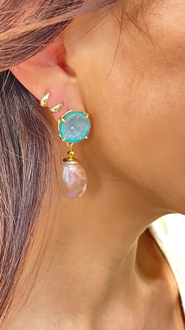 Close-up of a person's ear adorned with stylish jewelry. The ear features an Aria V Aria Earrings gold cartilage cuff and drop earring with a sparkly blue stone set in gold plated brass at the top, followed by a faceted pink teardrop gemstone. The person's hair is partially visible, with a light brown hue.