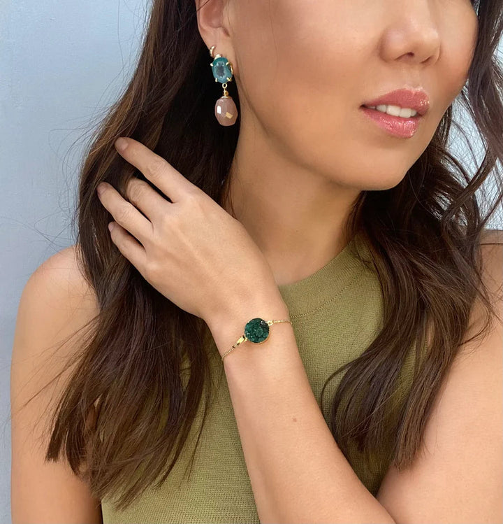 A woman with long brown hair wears a sleeveless, green top and accessorizes with a green stone bracelet and gold plated brass Aria Earrings by Aria V featuring green and pink stones. Her hand is gently placed near her collarbone, highlighting the jewelry against her skin.