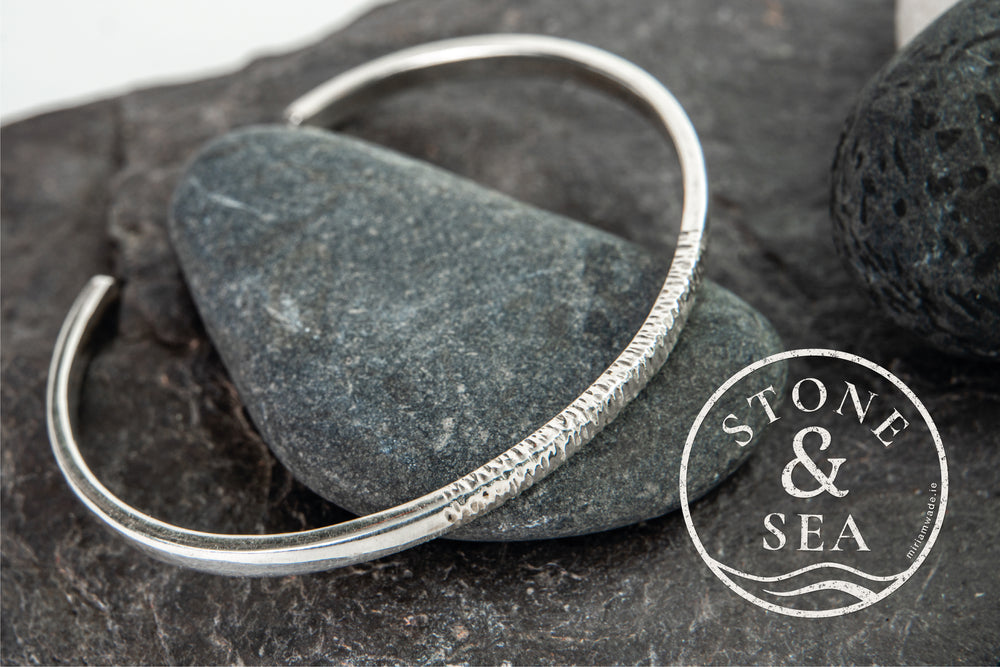 A textured Stone & Sea Silver Bangle by Miriam Wade rests on smooth, dark stones, inspired by organic forms along the Irish coastline. The "STONE & SEA" logo with wave graphics appears in the bottom right corner.