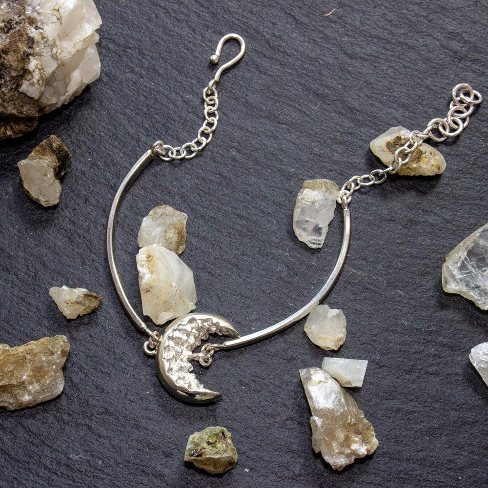 An Aud At Piece Bracelet Silver featuring a crescent moon charm is displayed on a dark slate surface, surrounded by scattered quartz crystal chunks. The moon charm has tiny star details, and the bracelet includes an adjustable chain with a hook clasp.