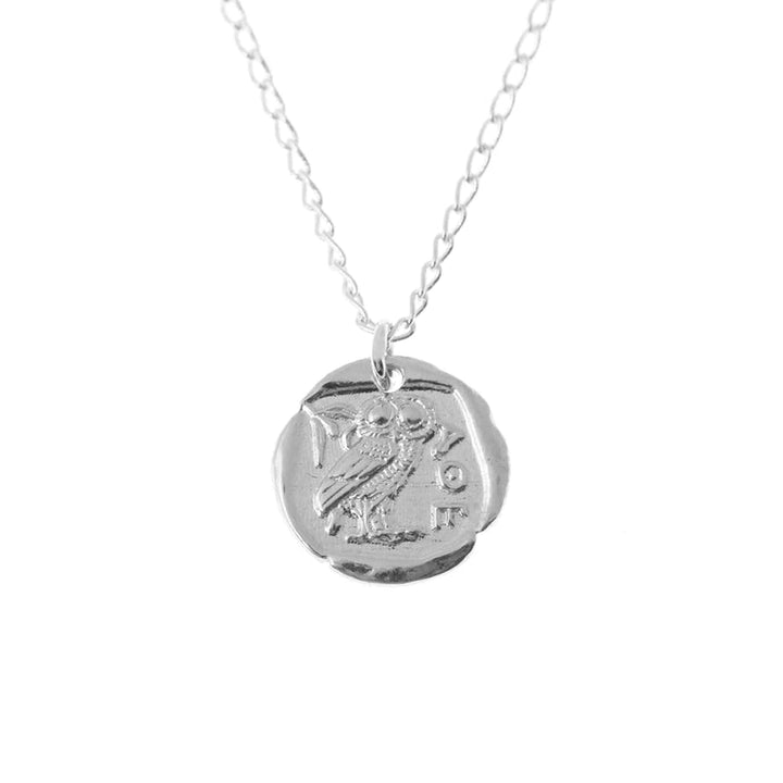 A sterling silver jewelry piece, the Andrea Mears Jewellery Athena Coin Necklace Silver features a round pendant with the embossed design of an owl and Greek letters. The pendant has a slightly irregular, ancient coin-like appearance, reminiscent of an Athena goddess pendant, and is connected to a delicate silver chain.