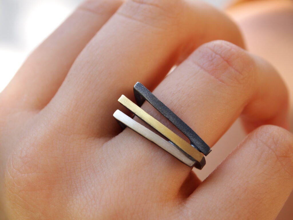 A hand wearing the MarCucurella Athens Silver & Gold Ring, consisting of three parallel, rectangular bands in oxidized silver, 18k gold plate, and black shades. The bands are stacked closely together, creating a sleek and contemporary look. The background is blurred.