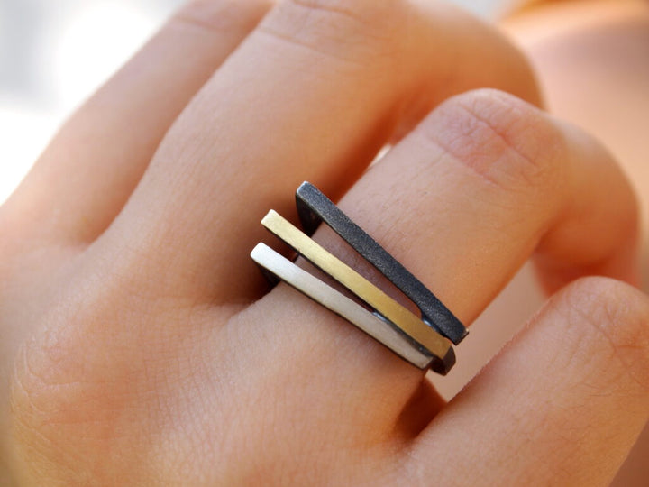 A hand wearing the MarCucurella Athens Silver & Gold Ring, consisting of three parallel, rectangular bands in oxidized silver, 18k gold plate, and black shades. The bands are stacked closely together, creating a sleek and contemporary look. The background is blurred.