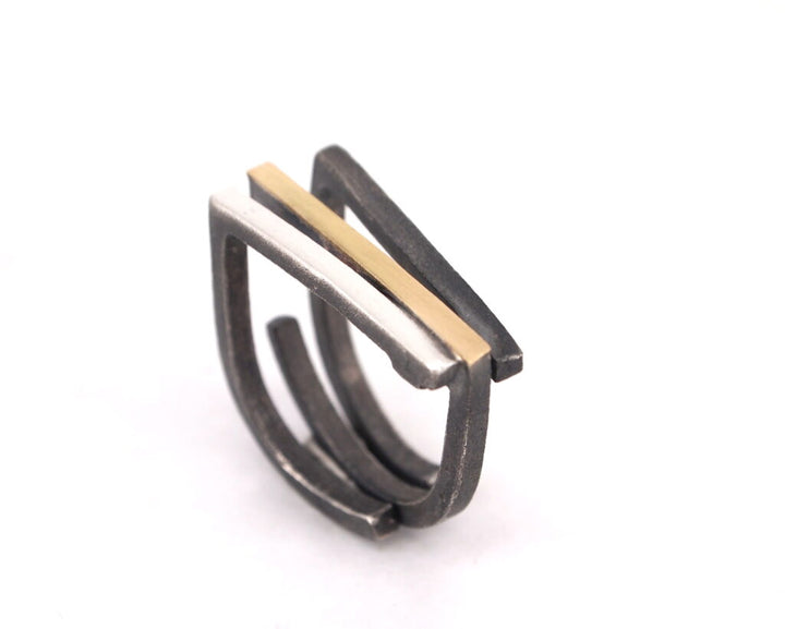 A modern, minimalist handmade ring featuring a stacked design with two open loops. Made of oxidized silver, the Athens Silver & Gold Ring by MarCucurella has a section in the center that is 18k gold plate. The overall aesthetic is sleek and geometric. The background is plain white.