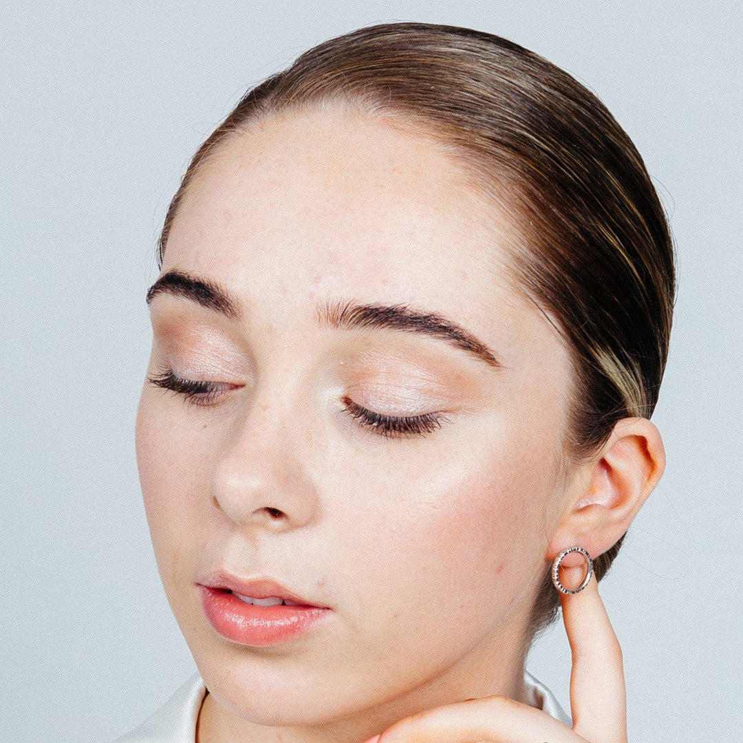 A person with slicked-back hair is shown from the shoulders up, slightly facing downwards with closed eyelids. They have light makeup and are touching their Into The Light Circle Earrings Silver by Aud with one hand. The background is plain and neutral, emphasizing the elegant texture detail of their accessories.