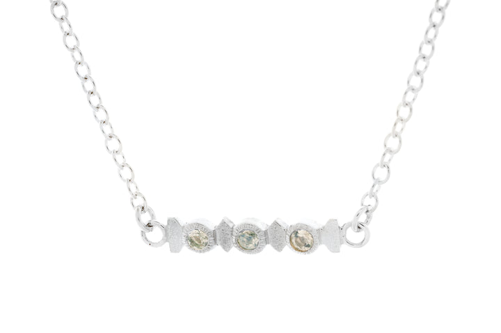 The Aud Midnight Necklace is a minimalistic and elegant piece featuring a delicate Sterling Silver chain and a horizontal bar pendant adorned with three small, round, faceted moonstones.