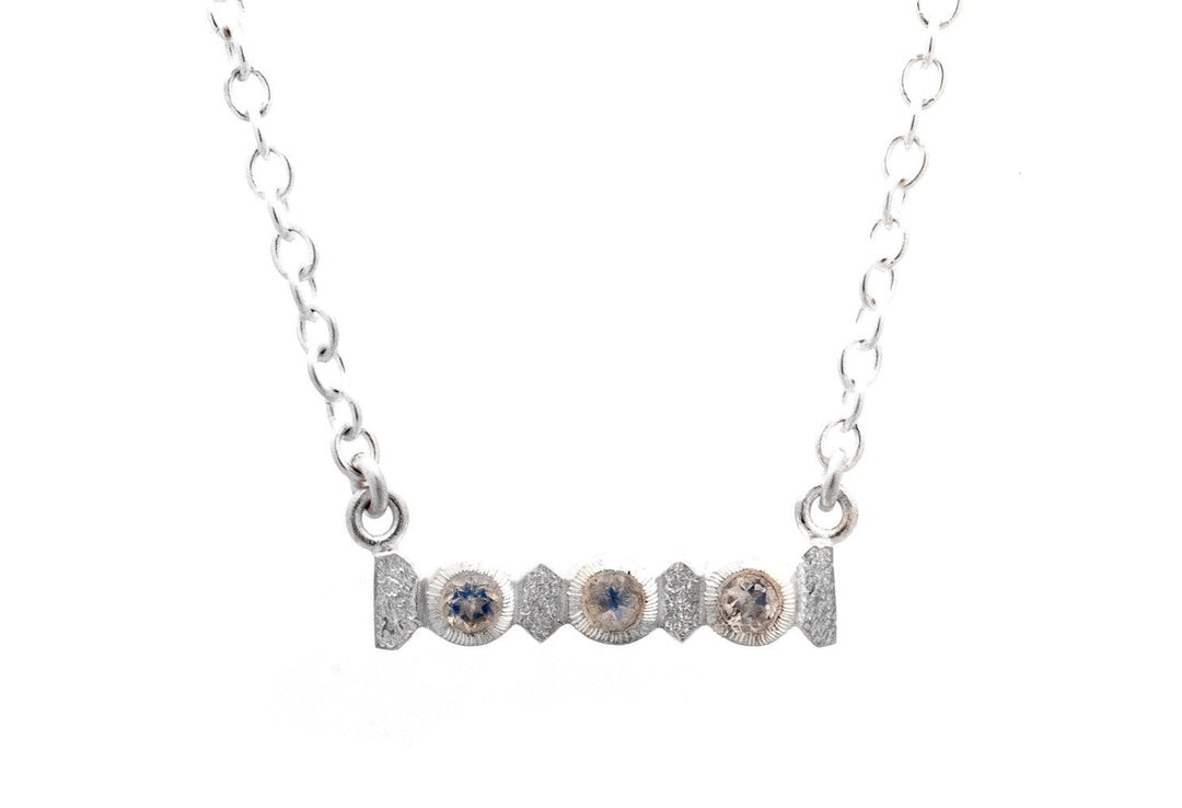 A silver necklace featuring a horizontal bar pendant adorned with a pattern of five round, blue and white faceted moonstones. Dubbed the Midnight Necklace by Aud, the pendant is attached to a delicate Sterling Silver chain with oval links, giving it a modern and elegant look. The background is plain white.