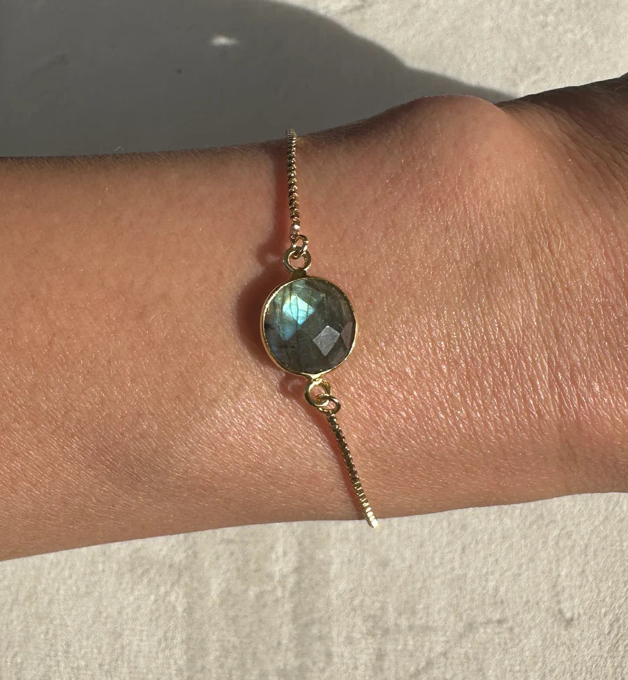 The Aurora Single Drop Bracelet by Aria V features a gold-plated design with a round, faceted labradorite stone that glimmers beautifully on the wrist in natural light. Handmade in Dublin, this elegant piece adds artisanal charm to any ensemble.