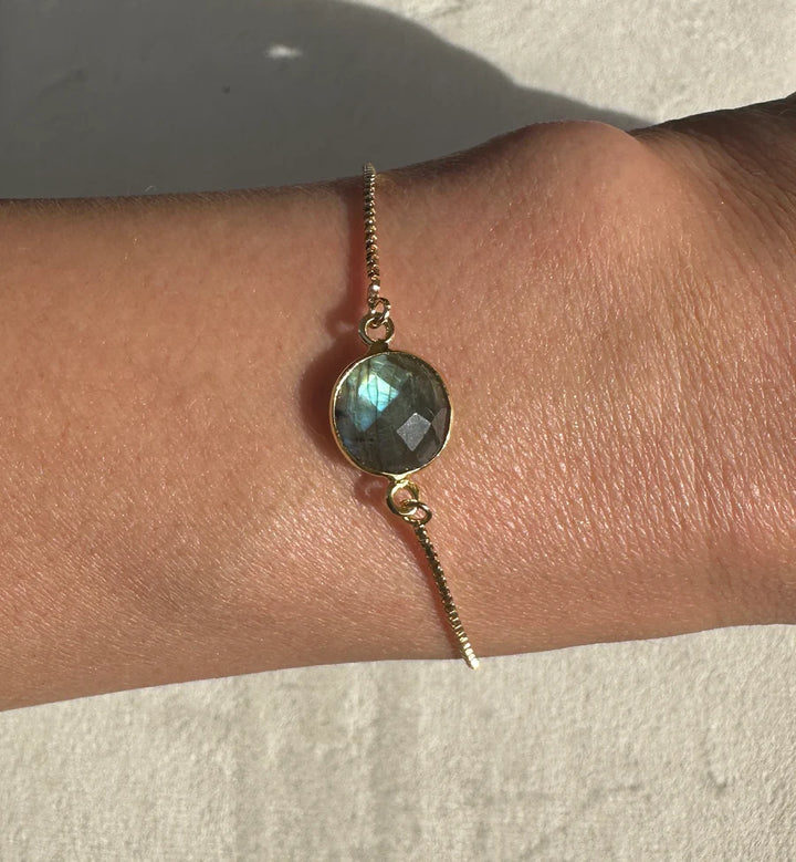 The Aurora Single Drop Bracelet by Aria V features a gold-plated design with a round, faceted labradorite stone that glimmers beautifully on the wrist in natural light. Handmade in Dublin, this elegant piece adds artisanal charm to any ensemble.