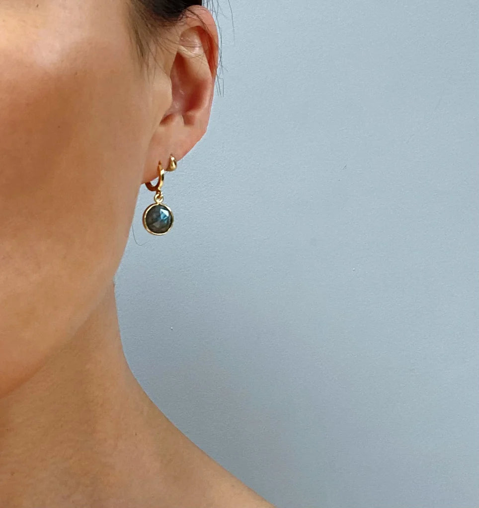 Close-up of a person's ear wearing Aria V's Aurora Earrings—a small gold hoop handcrafted earring with a round, dark stone pendant. The face is partially visible against a plain light gray background.