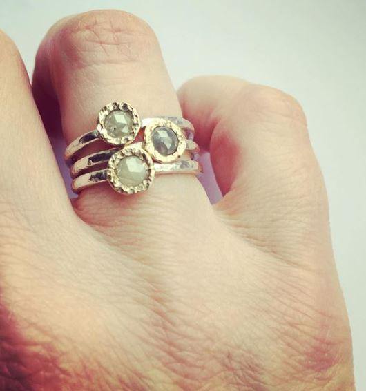 A close-up image of a hand wearing three sterling silver stacking rings on the middle finger, each ring featuring a faceted, circular gemstone. One ring showcases an **Abbie Dixon Aurous Ring Silver Grey Diamond** set in a hammered finish band. The background is plain and light in color, highlighting the rings.